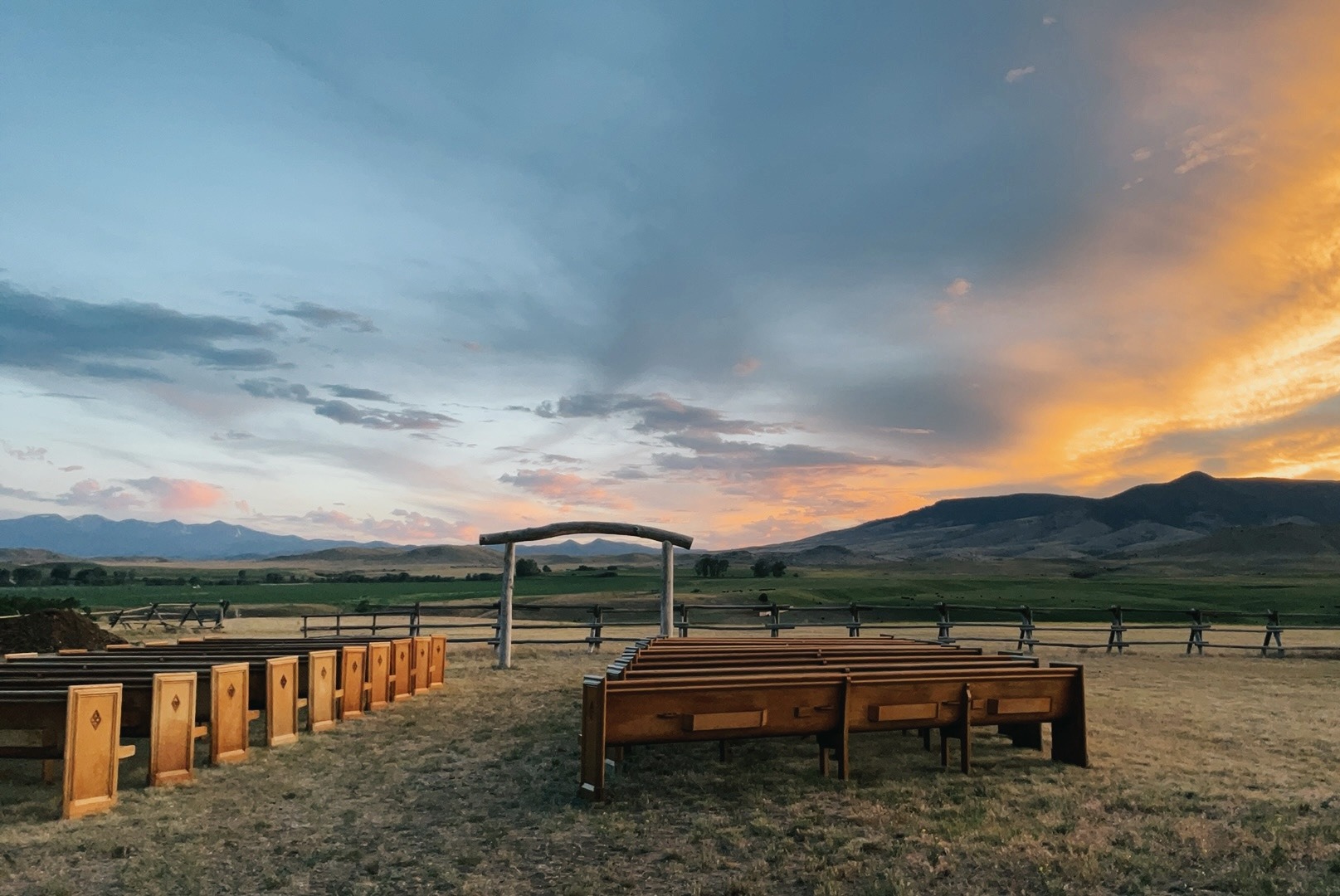 Wooden pews facing the handmade ceremony arch in the meadow at sunset, mountains behind