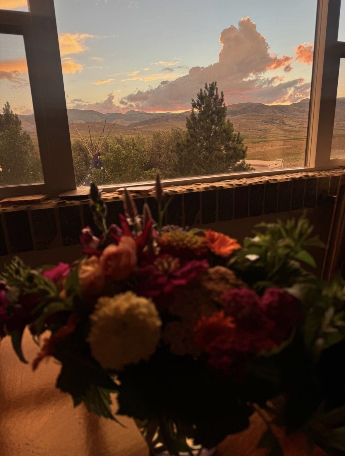 A farm bouquet on the windowsill at sunset, the tipi, pines and mountains glowing beyond the glass
